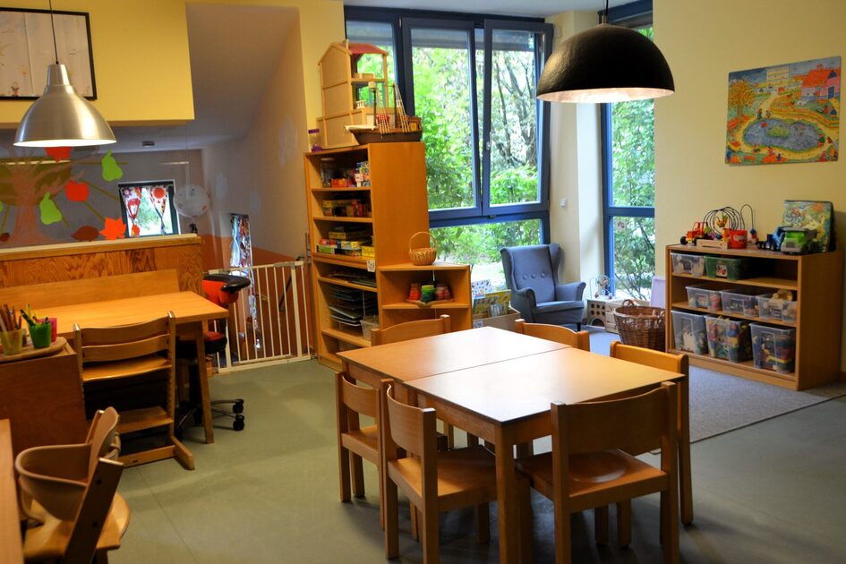 Ein Gruppenraum der Kindertagesstätte mit Tischen und Stühlen. Im Hintergrund der Teppich der Bauecke