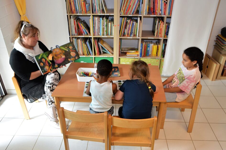 Die Bücherei. An einem Tisch sitzen drei Kinder mit einer Erzieherin, die gemeinsam ein Buch anschauen. An der Wand steht ein großes Bücherregal