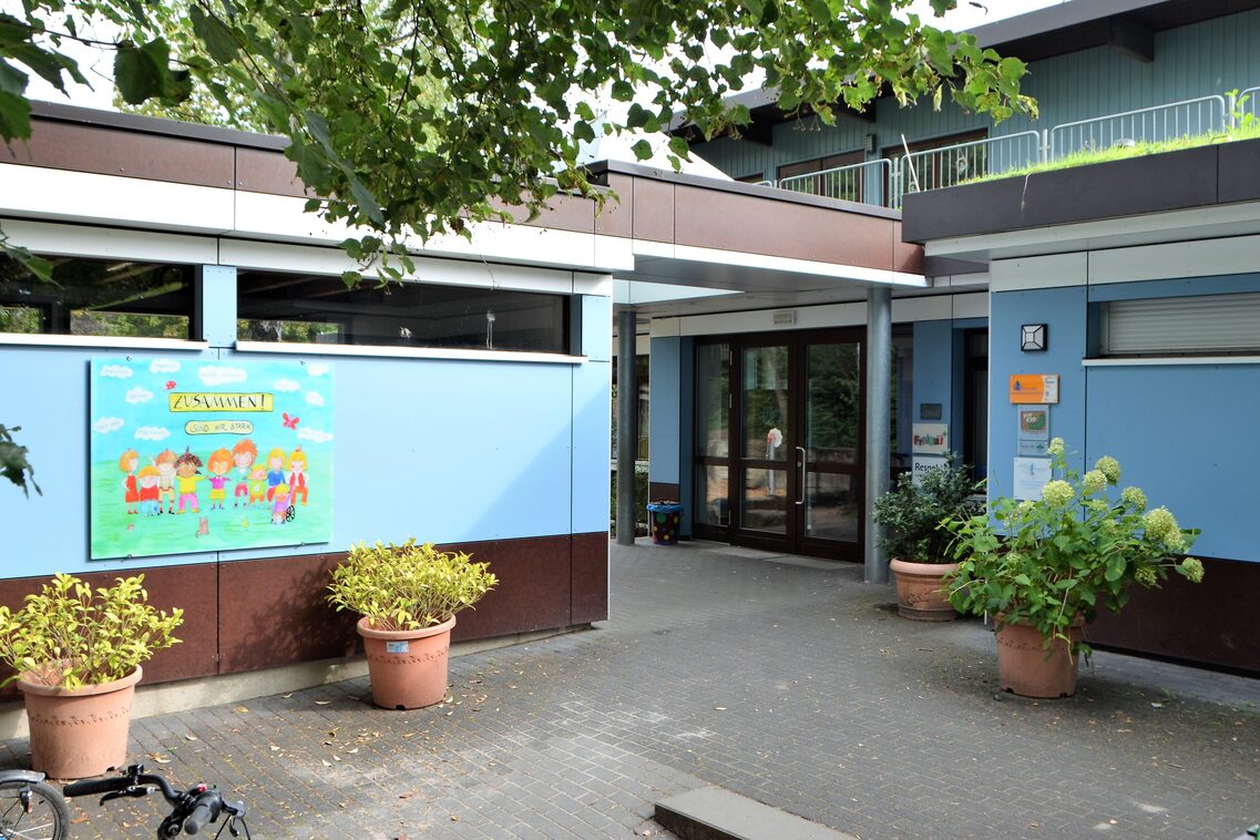 Der Eingangsbereich der Kindertagestätte mit Pflanzkübeln. An der rechten Wand des Gebäudes hängt ein Bild auf dem Kinder gemalt sind