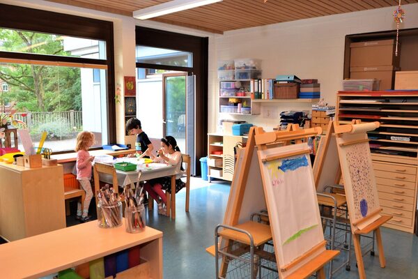 The art studio with shelves for paper and paints. Three children are painting at a table. In the foreground are two easels with pictures