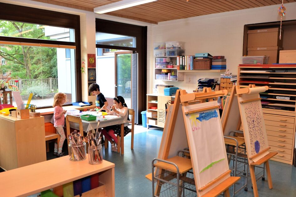 Das Kunstatelier mit Regalen für Papier und Farben. An einem Tisch sitzen drei Kinder beim malen. Im Vordergrund stehen zwei Staffeleien mit Bildern