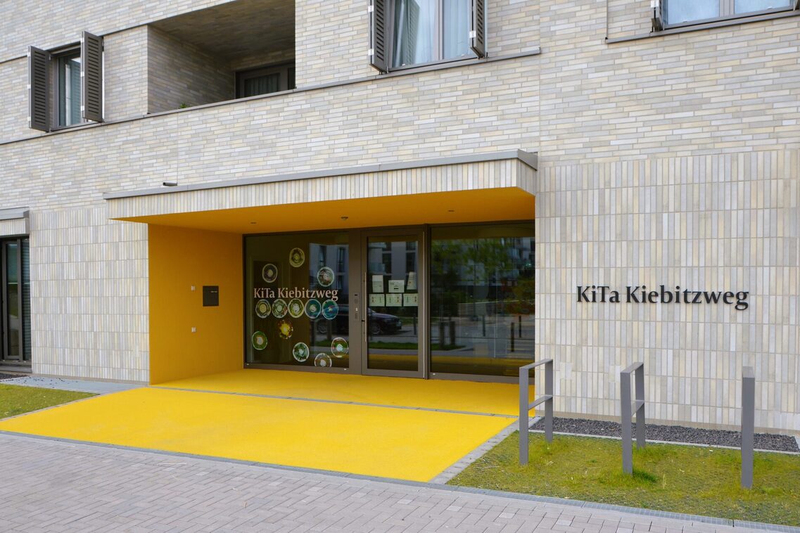 Der Eingang der Kindertagesstätte. Die Glastür ist unter einer gelben Überdachung. Rechts stehen Fahrradständer
