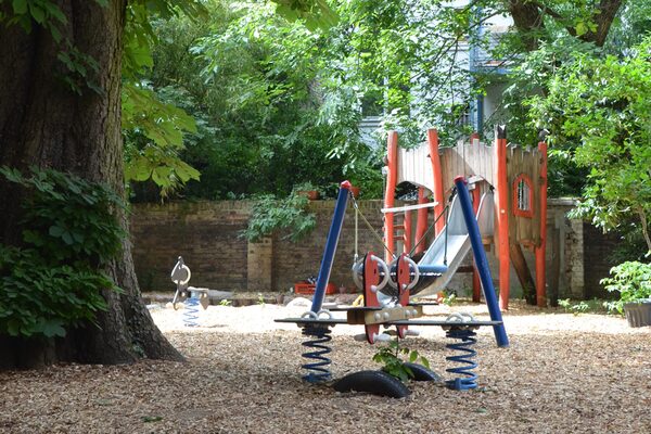 Das Außengelände der Kindertagesstätte. Im Hintergrund steht ein Kletterturm aus Holz mit einer Rutsche. Links ist ein großer Baum