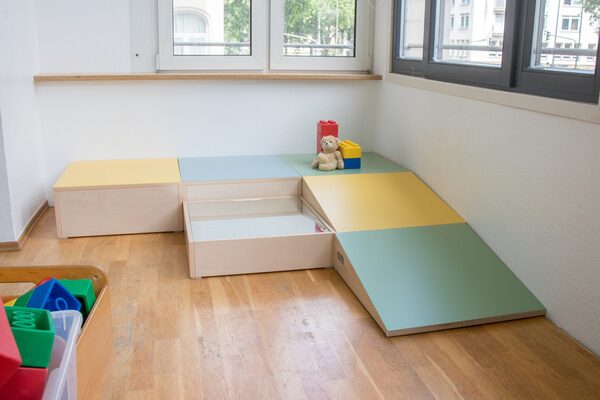 Der Spielbereich der Krippengruppe. In der Ecke stehen Holzpodest mit einer schrägen Ebene. Links im Vordergrund eine Kiste mit Bausteinen