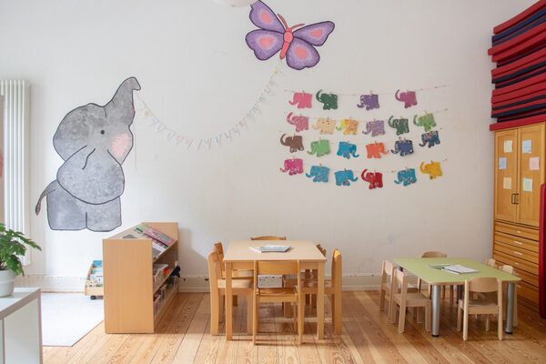 Ein gruppenraum im Kindergarten. Im Raum stehen Tische und Stühle. An der Wand hängt ein großer Elefant, der an einer Schnur einen Schmetterling am Rüssel hält.