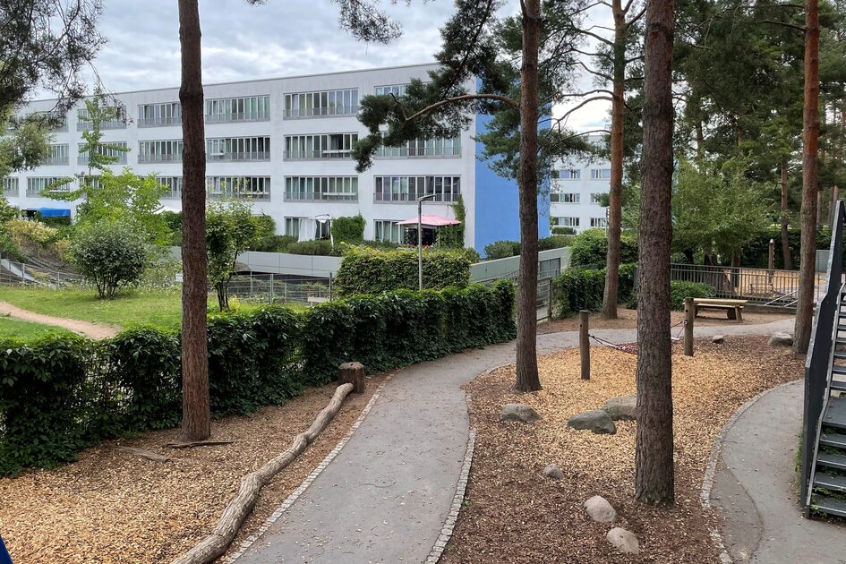 Das Außengelände der Kindertagesstätte mit großen Bäumen und einem Weg. Das Gebäude steht auf der rechten Seite - links ist eine Hecke. Eine Treppe führt zum Außengelände