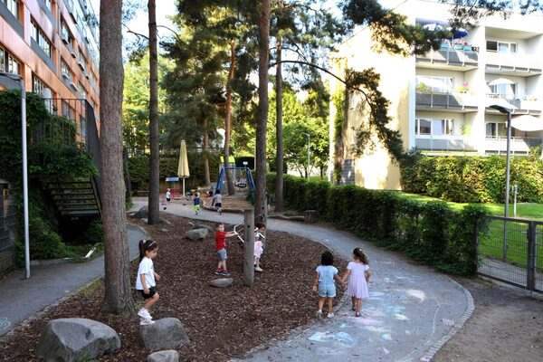 The outdoor area of the daycare center with a curved path between trees and a hedge. In the background, a sand play area with a swing
