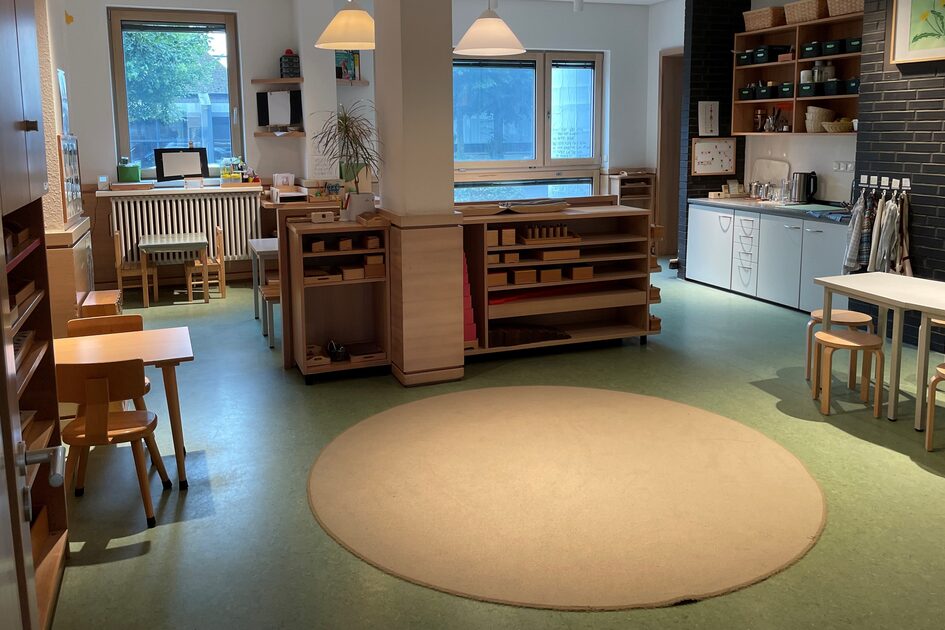 Ein Gruppenraum der Kindertagesstätte. Im Vordergrund liegt ein beiger runder Teppich. Dahinter stehen Regale mit Beschäftigungsmaterial. Vor den Fenstern stehen Tische