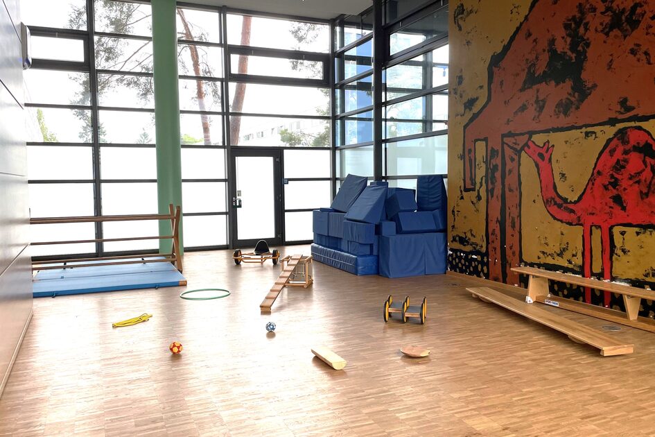 Der Bewegungsraum mit einer großen Fensterfront. Im Vordergrund sind eine Leiter, Bälle, ein Kippelbrett und ein Pedalo auf dem Boden. Rechts an der Wand ist eine Wandgemälde mit einem Kamel. DAvor stehen große Matten