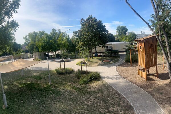 Das Außengelände der Kindertagesstätte. Rechts steht ein Baum mit einem Holz-Blockhaus. Durch das Gelände schlängelt sich ein Weg.