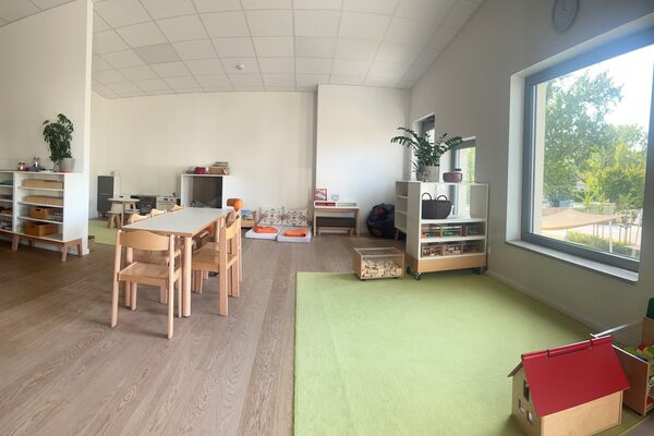 Vor dem Fenster an der rechten Seite liegt ein grüner Teppich. Davor steht ein Tisch mit Stühlen. Im Hintergrund sind Regale mit Spielmaterial