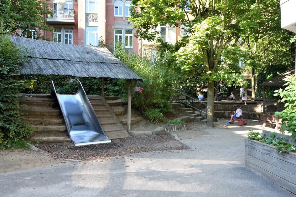 das Außengelände der Kindertagesstätte mit einer breiten Rutsche an einem Häuschen. Auf der rechten Seite stehen Hochbeete