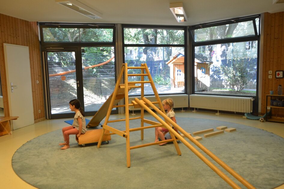 Der Bewegungsraum der Kindertagesstätte mit einer großen Fensterfront. In der Mitte ein großer runder Teppich mit einer Leiter und Balancierstangen