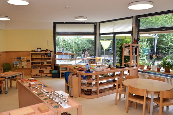 A group room with a large window front overlooking the outside area. There are tables with chairs and various shelves with play materials in the room