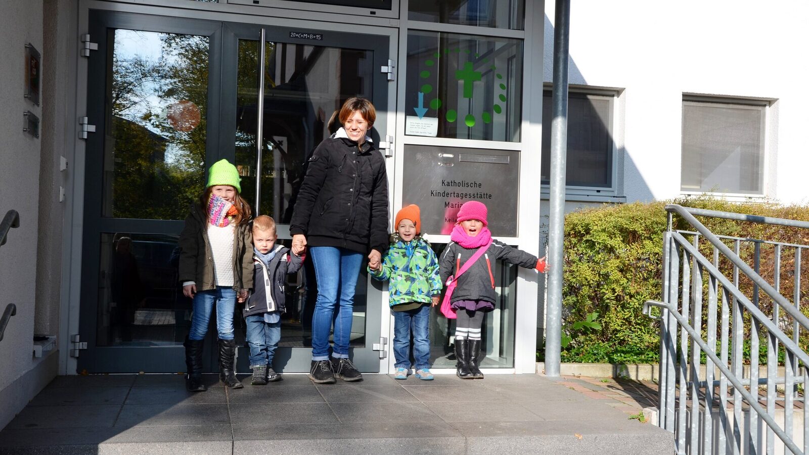 Man sieht den Eingang der Kita. Eine Frau mit Kindern an der Hand steht vor der Tür