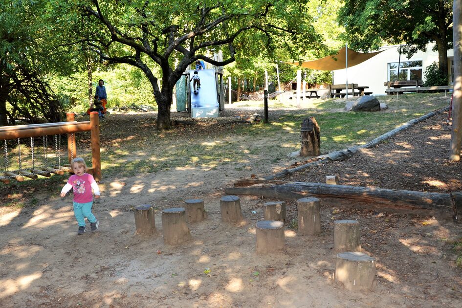 Das Außengelände der Kindertagesstätte. Am rechten Bildrand die Holzumrandung für das Klettergerüst - links eine Hängebrücke. Im Hintergrund ist eine Rutsche