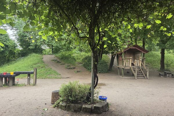 The outdoor area of the daycare center. There is a tree in the center of the picture. In the background on the left is a waterhole, on the right is a wooden playhouse with stairs