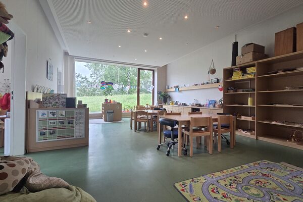The group room with a large window facing the outside area. On the right is a large shelf with play materials, in front of which are two tables with chairs.