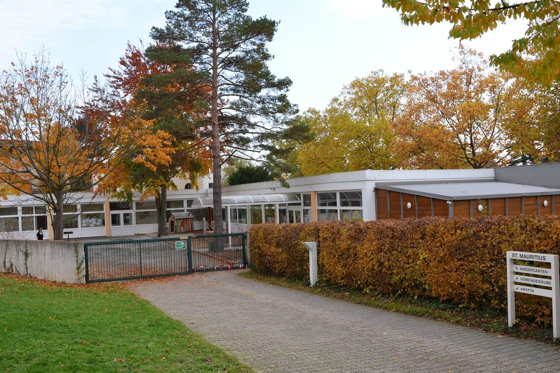 Zugang zur Kindertagesstätte mit dem Gebäude im Hintergrund. Rechts ist eine Hecke - davor steht das Schild der Kindertagesstätte