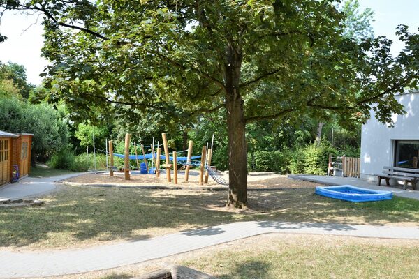 Das Außengelände der kindertagesstätte. In der Mitte steht ein großer Baum, dahinter ein Kletterelement. neben dem baum ist ein Plantschbecken