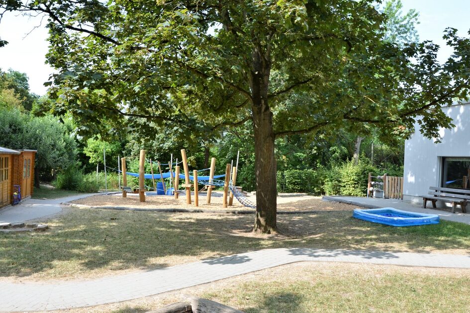 Das Außengelände der kindertagesstätte. In der Mitte steht ein großer Baum, dahinter ein Kletterelement. neben dem baum ist ein Plantschbecken