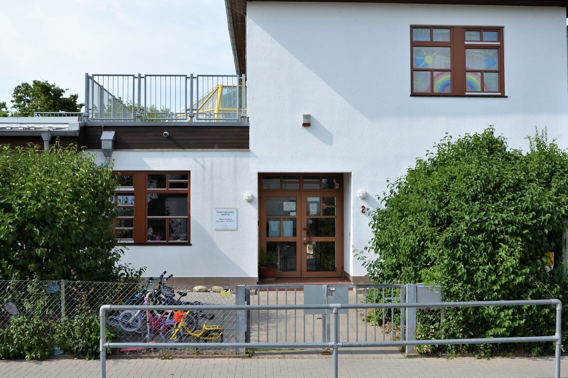 Der Eingangbereich der Kindertagesstätte mit Büschen auf der rechten und linken Seite. Hinter dem Zaun stehen Fahrräder