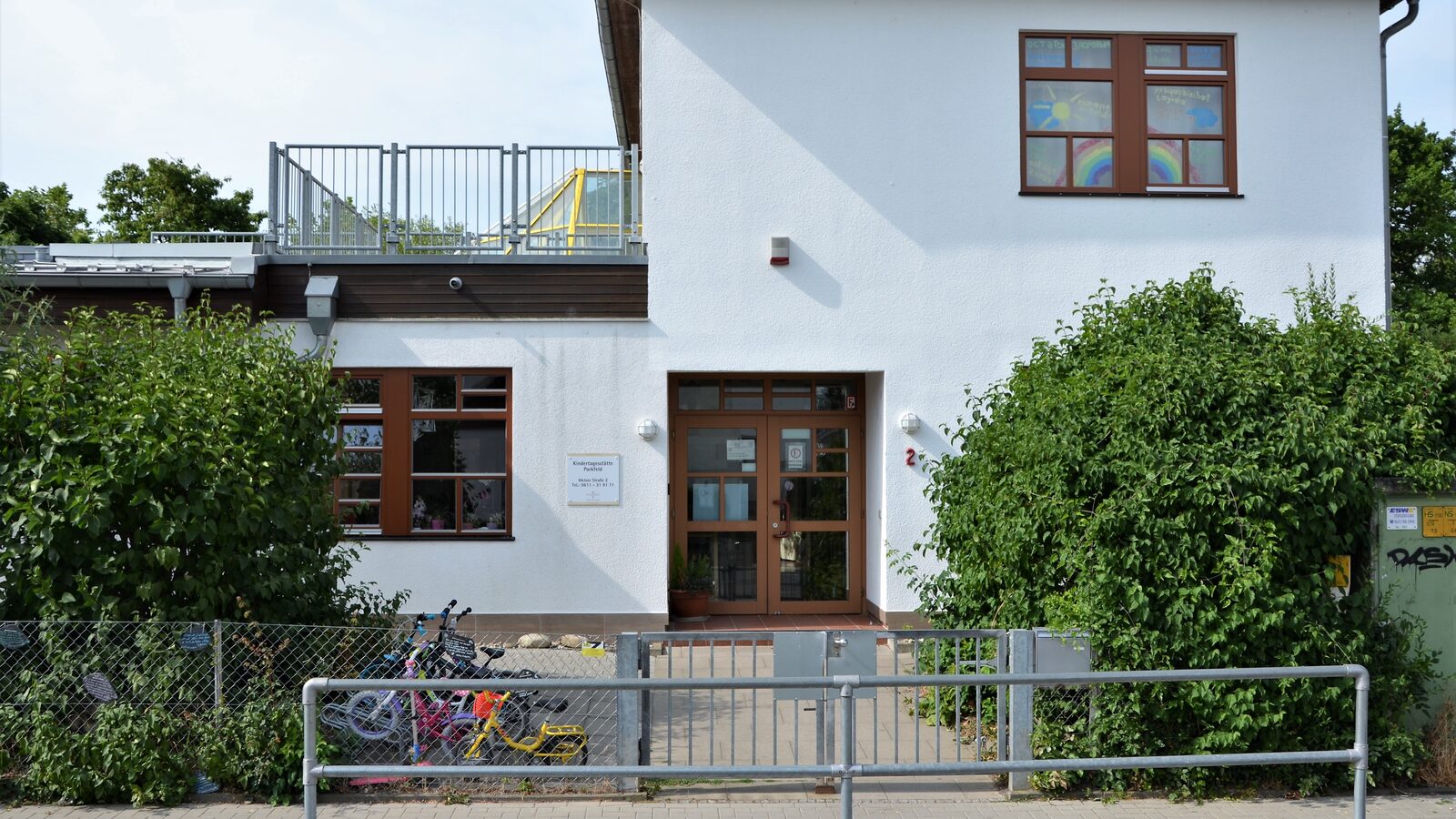 Der Eingangbereich der Kindertagesstätte mit Büschen auf der rechten und linken Seite. Hinter dem Zaun stehen Fahrräder