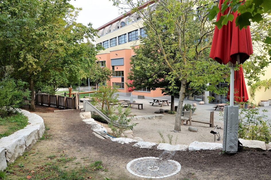 Das Außengelände mit der Kindertagesstätte im Hintergrund. Links ist eine Rutsche und rechts steht ein roter, zusammengeklappter Sonnenschirm.