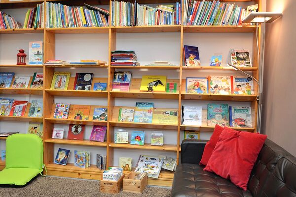 The library of the daycare center with a large shelf full of children's books. There is a brown sofa on the right-hand side.