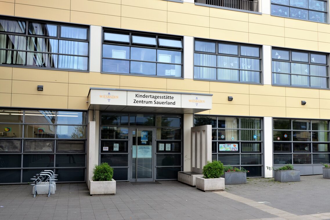 Der Eingang der Kindertagesstätte mit großen Fenstern auf der rechten und linken Seite. Vor der Haus stehen Pflanzkübel und ein Fahrradständer