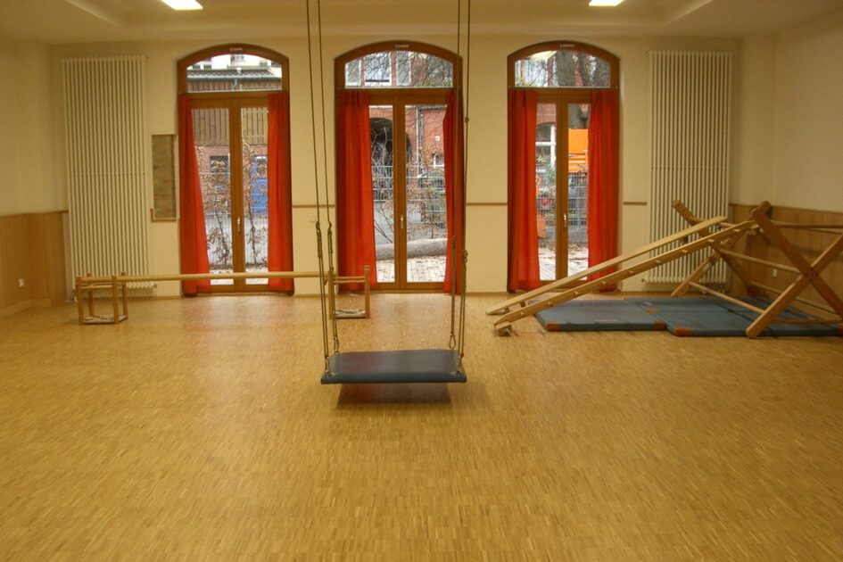 Der große Bewegungsraum mit Kletterelementen und einer Hängeschaukel an der Decke. Die Fenster mit rotem Rahmen zeigen zum Außengelände