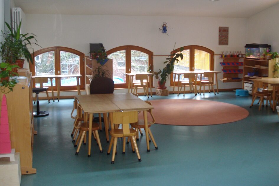 Der Gruppenraum mit Tischen und Stühlen. Auf dem Boden liegt ein runder Teppich. An den Wänden stehen Regale mit Spielmaterial