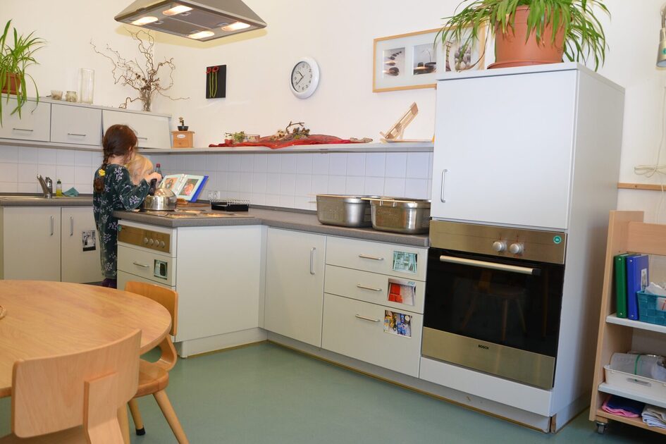 Die Kinderküche mit einer Küchenzeile mit Backofen, Herd, Kühlschrank und Spüle. Ein Kind steht am Herd auf dem ein Kessel steht.