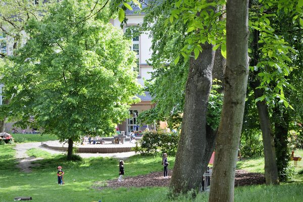 Die Kindertagesstätte liegt in einem großen Park mit hohen alten Bäumen. Im Bildhintergrund ist das Gebäude zu sehen.