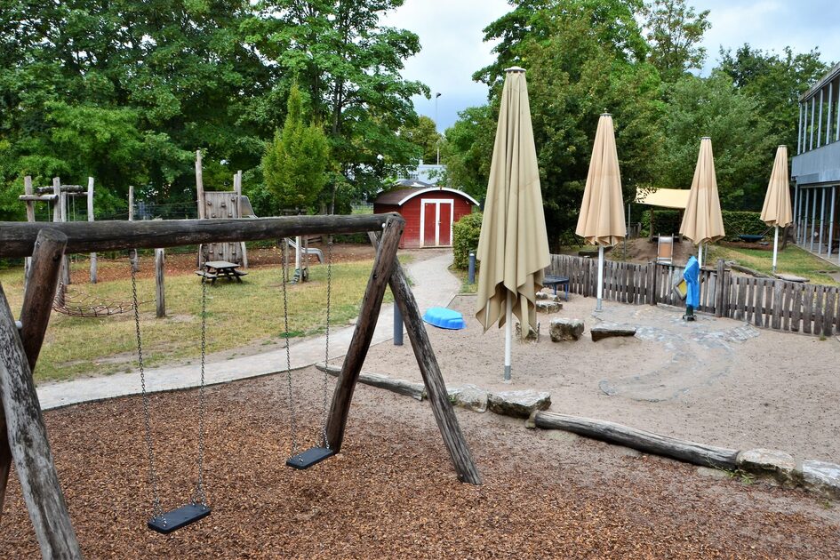 Das Außengelände der Kindertagesstätte. Im Vordergrund eine Schaukel in einem abgegrenzten Bereich mit Rindenmulch Im Sand stehen zwei große weiße Schirme
