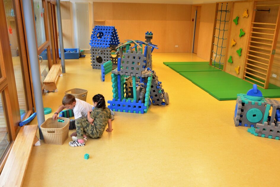 Zwei Kinder spielen im Bewegungsraum mit großen Steckelementen, die zu Häusern zusammengesteckt werden können. An der rechten Seite ein Klettergerüst an der Wand