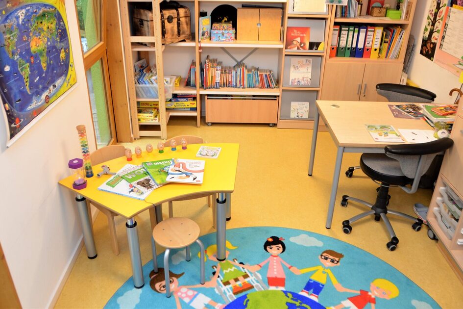 Die Bücherei mit zwei Tischen zum lesen und arbeiten. Auf dem Boden liegt ein runder Teppich. An der Wand steht ein großes Regal mit Büchern und Ordner