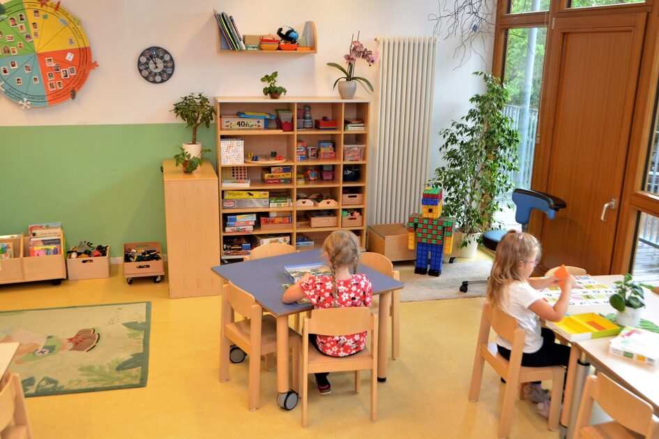 Eine Gruppe im Elementarbereich. An zwei Tischen sitzen Kinder und spielen. Auf dem Boden liegt ein Teppich und an der Wand ist ein Regal mit Spielmaterialen.
