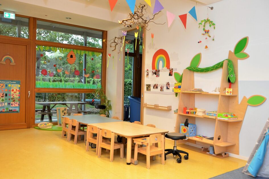 Ein Gruppenraum in der Krippe mit viel Freifläche auf dem Boden zum krabbeln. Auf der rechten Seite stehen Tische mit kleinen Stühlen. An der Wand ist ein Regal und ein Regenbogen aus Papier.