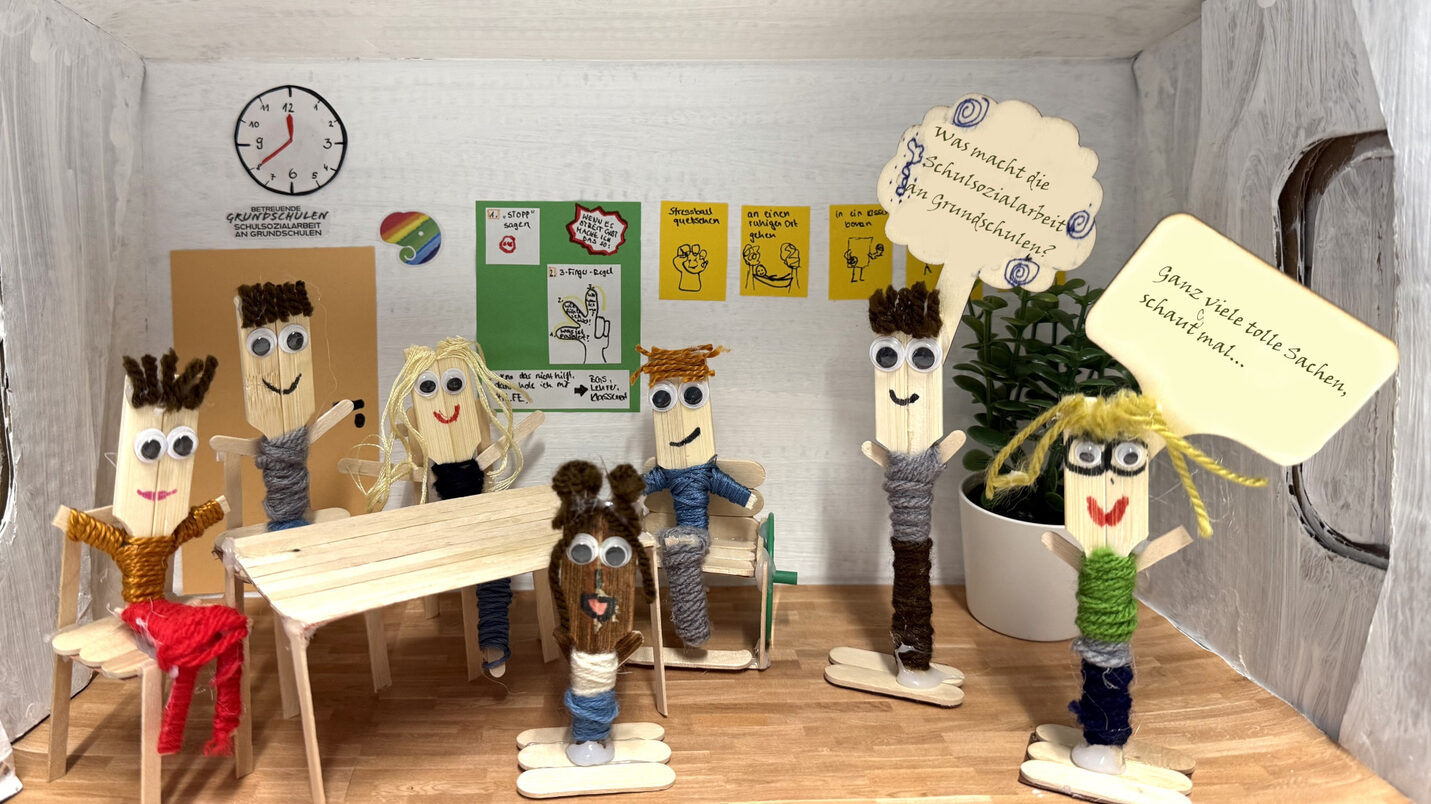 Gebastelter Schuhkarton als Klassenzimmer in dem sieben aus Holz und Wolle gebastelte Figuren sind. Fünf Kinder sitzen um einen Tisch herum. Ein stehendes Kind hält ein Schild hoch auf dem steht "Was macht die Schulsozialarbeit an Grundschulen?" Die Schulsozialarbeiterin rechts neben dem Kind hat auf ihrem Schild "Ganz viele tolle Sachen, schaut mal..." geschrieben.