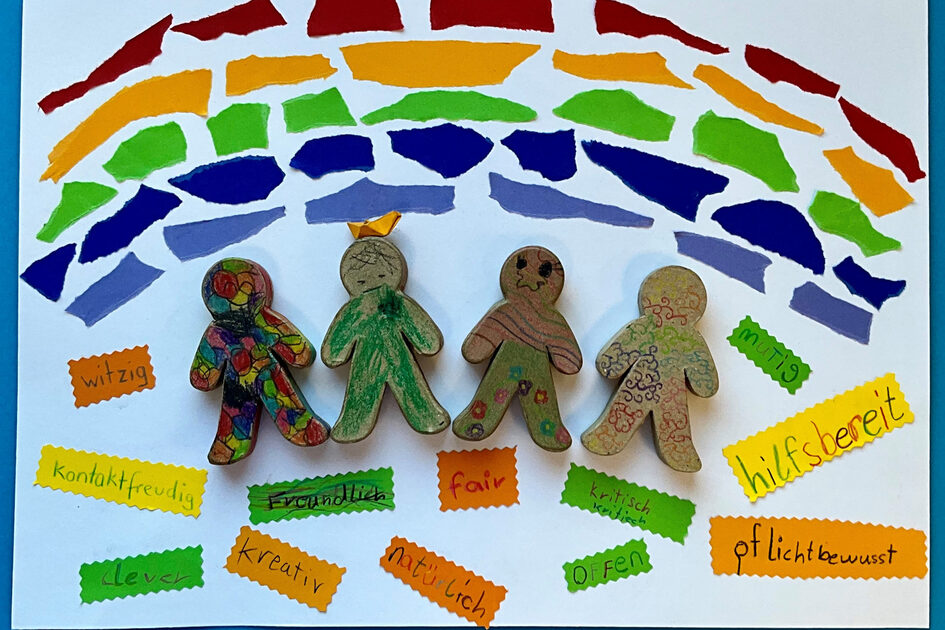 Gebasteltes Bild mit vier Holzfiguren, die unter einem Regenbogen stehen. Darunter stehen auf Papier geschrieben verschiedene Eigenschaften wie witzig, kontaktfreudig, clever, kreativ, freundlich, natürlich, fair, offen, kritisch, pflichtbewusst, hilfsbereit.