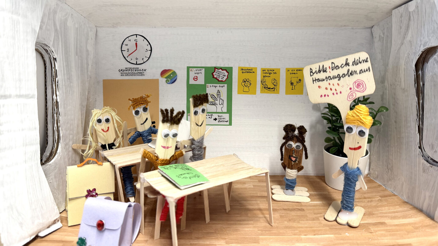 Gebastelter Schuhkarton als Klassenraum. Darin sitzen vier aus Holzessstäbchen gebastelte Kinder an ihrem Tisch und hören der Lehrerin zu. Ein Kind steht neben der Lehrerin, die ein Schild hochhält auf dem steht: "Bitte pack deine Hausaufgaben aus"