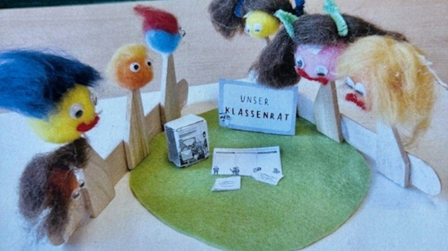 Фігури з дерева та фетру сидять навколо зеленого килима з табличкою "Наша класна рада", поштовою скринькою та записками.