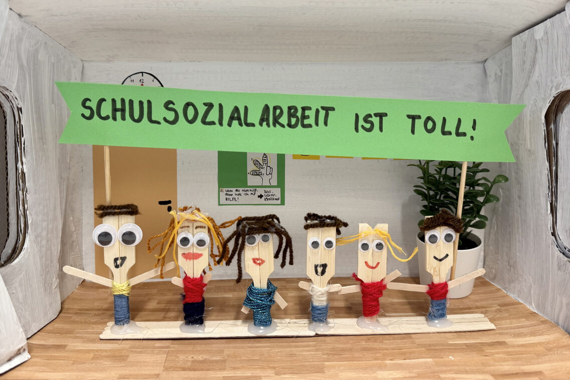 Gebastelter Schuhkarton als Klassenraum. Sechs aus Holzstäbchen gebastelte Figuren mit großen Kulleraugen und Haaren aus gelber und brauner Wolle stehen in einer Reihe nebeneinander. Sie halten ein grünes Schild hoch, auf dem steht "Schulsozialarbeit ist toll!"