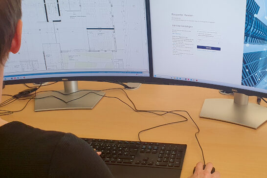 zwei Computerbildschirme von denen einer eine technsiche Zeichnung eines Gebäudegrundrisses und der andere die Startseite des hessischen Bauprotals zeigt. Davor eine Person von hinten, angeschnitten sodass nur ein Teil des Hinterkopfes, eine Hand, die eine Computermaus hält zu sehen ist und die vor sich eine Computertatstatur auf dem Schreibtisch hat.