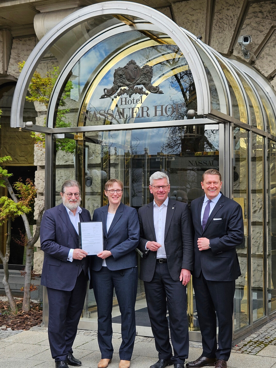 امرأة وثلاثة رجال يرتدون بدلات عمل يقفون بجانب بعضهم البعض أمام مدخل فندق ناساور هوف في فيسبادن ويبتسمون للكاميرا.