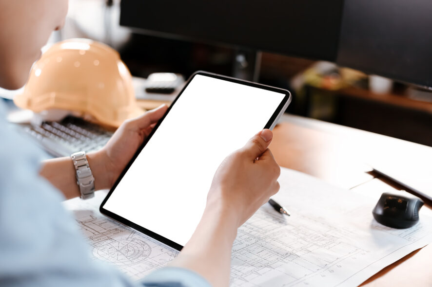 Ένα άτομο κρατά ένα tablet στα χέρια του, ένα κράνος κατασκευής και ένα σχέδιο κατασκευής βρίσκονται σε ένα γραφείο στο βάθος.