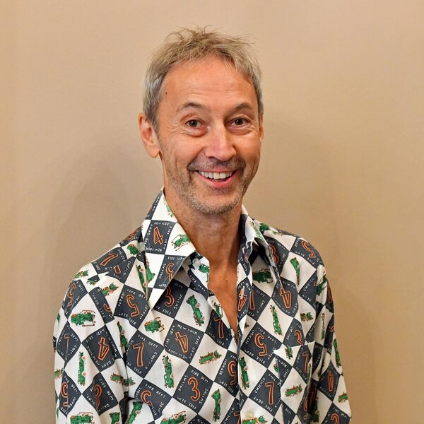 Мъж с посивяла коса носи риза с шарка от цифри и малки зелени фигури на бежов фон.