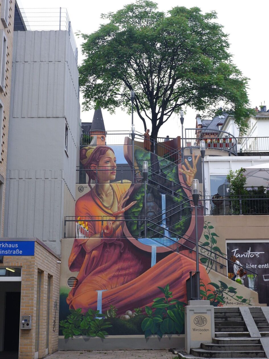 Wandgemälde eines künstlerischen Motivs mit einer weiblichen Figur, die einen Behälter mit Pflanzen und Wasser hält, umgeben von urbanem Umfeld und Treppen. Oberhalb der Treppe steht ein Baum, es sieht aus als würde er aus dem Bild herauswachsen.