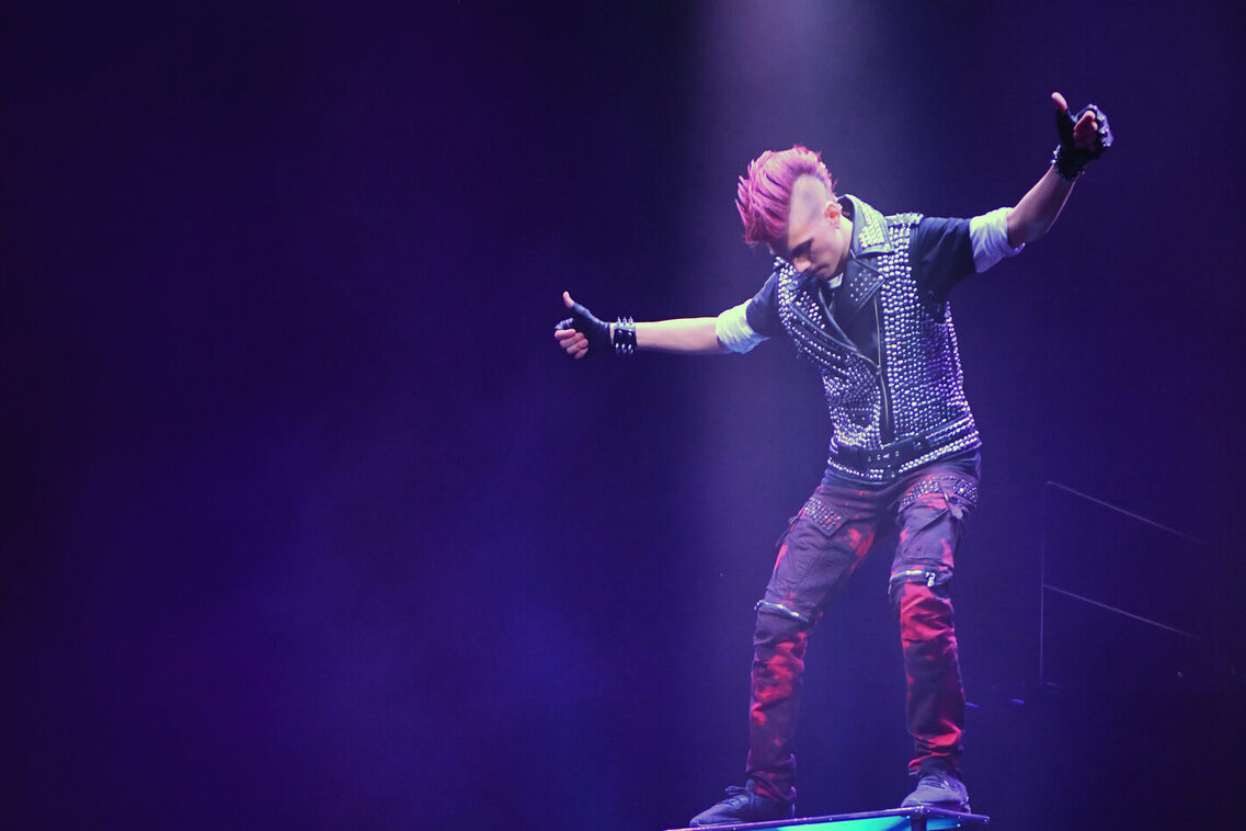 Artist Kaya Janeček balanciert auf einem Podest stehend auf einem Rola-Rola auf dem wiederum sieben aufeinandergestapelte Bretter stehen